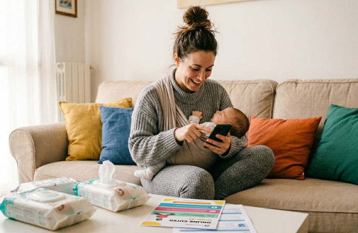 Mamma con bambino in fascia punta il dito sullo schermo dello smartphone che mostra un post di Lofacciodigital intitolato '3 tipi di clienti sei una Mamma Inconsapevole?'.